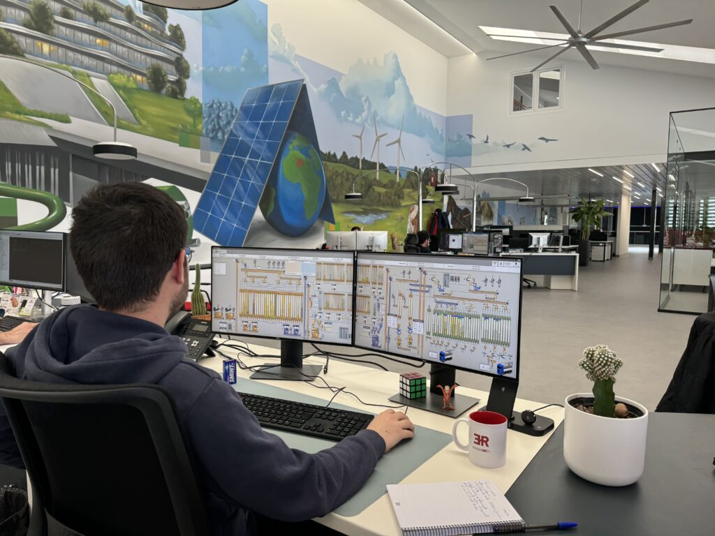 ER Ingeniería technician seen from behind, in front of two monitors showing a plant's process mimic diagrams, in a spacious office with a solar panel mural