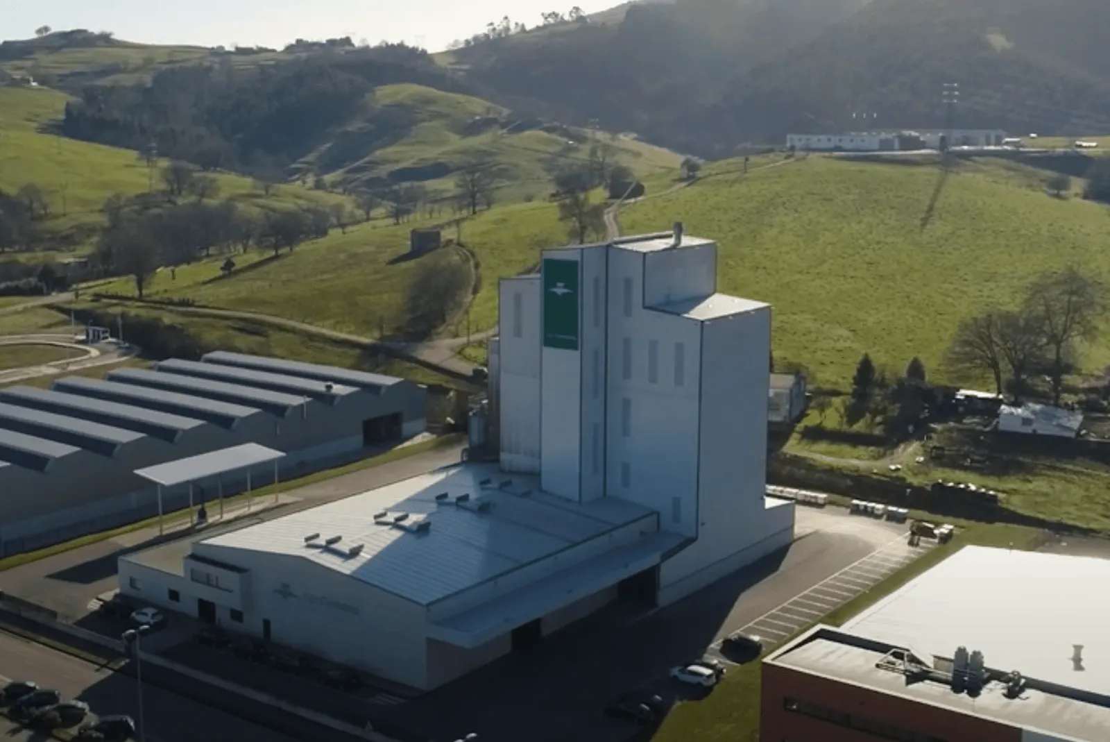 Aerial view of the Agrocantabria feed mill: white manufacturing tower with a green stripe and warehouses beside it, among meadows and hills