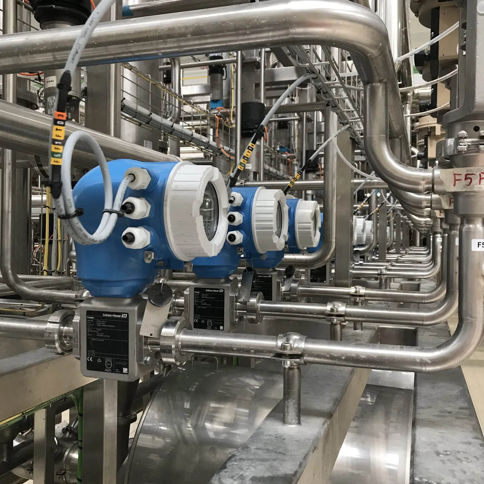 Flow meters with blue housings mounted on stainless steel pipes, with labelled wiring, in a process plant